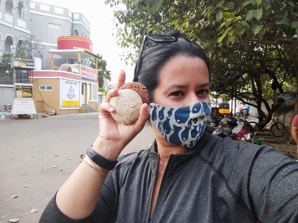 When solo stands on a quiet street in India, holding a piece of local street food while wearing a protective face mask. This image captures the essence of independent travel, cultural exploration, and mindful safety during solo journeys.